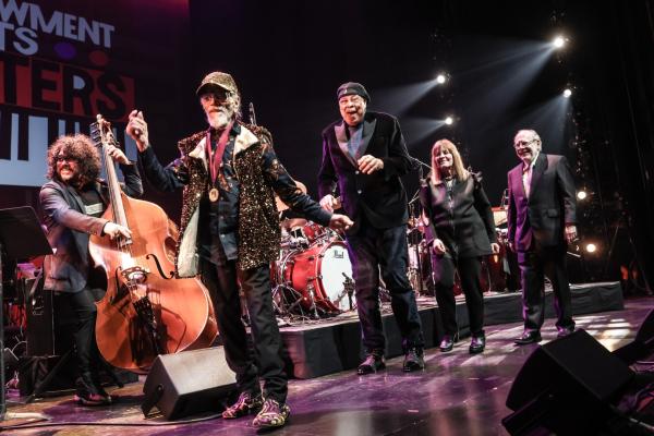 Three men and a woman smile and walk single-file onstage while musicians play behind them.