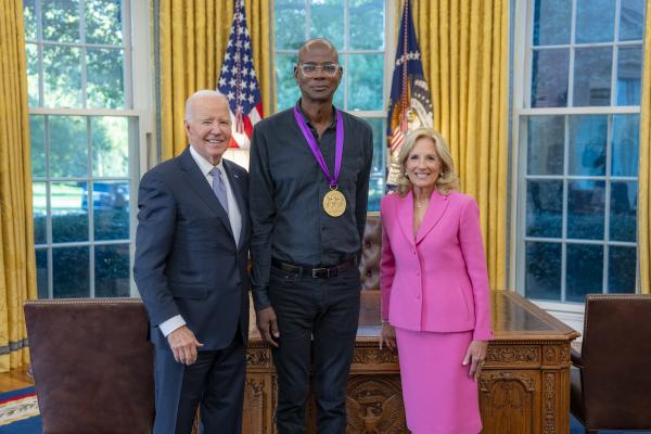 Tall bald Black man wearing glasses with a medal around his neck surrounded by older man wearing a blue suit and woman with blonde hair wearing a pink suit. 